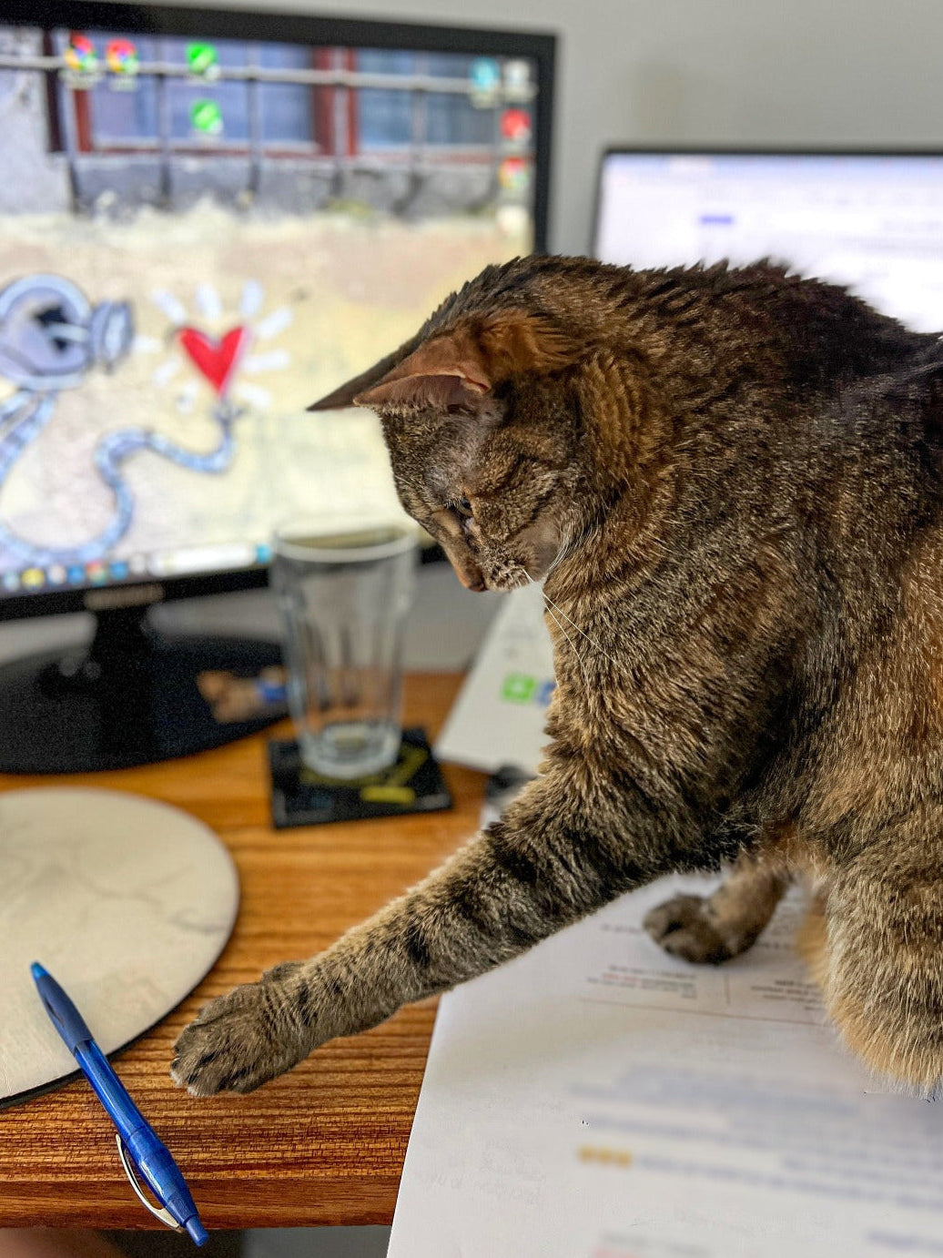 Cat sitting on a desk next to a computer monitor, playing with a pen.